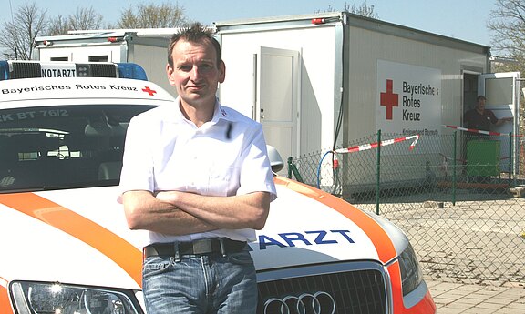 Ein Notarzt steht mit verschränkten Armen vor einem Rettungswagen des Bayerischen Roten Kreuzes. Im Hintergrund ist ein Gebäude mit dem Logo des Roten Kreuzes sichtbar. Es ist ein sonniger Tag.