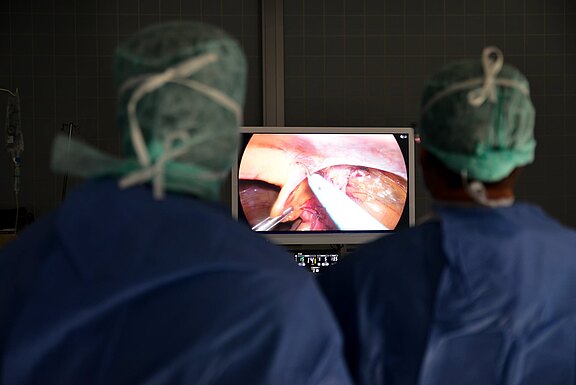 Zwei Chirurginnen/Chirurgen in OP-Kleidung betrachten einen Monitor, der eine laparoskopische Operation zeigt. Auf dem Bildschirm sind endoskopische Instrumente und Gewebe im Bauchraum zu sehen. Fokus auf Überwachung des Eingriffs durch das Team.