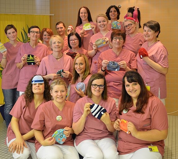 Gruppe von Pflegekräften in rosa Arbeitskleidung posiert in einem Klinikraum und hält bunte, selbstgemachte Babymützen in die Kamera; sie lächeln.