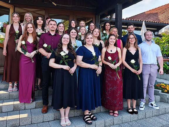 Gruppenfoto von rund 19 jungen Erwachsenen in festlicher Kleidung, auf Treppen posierend. Viele halten weiße Rosen und lächeln in die Kamera. Feierliche Stimmung, vermutlich Abschlussfeier oder Preisverleihung im Freien vor einem Gebäude.