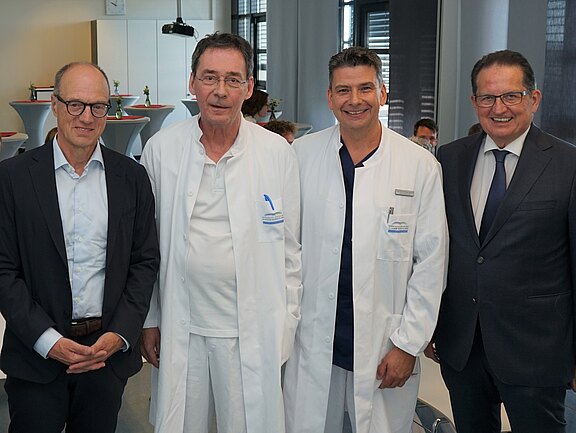 Fünf Personen stehen gemeinsam in einem Raum. Vier Männer tragen helle oder dunkle Anzüge sowie Arztkittel, während eine Frau in einem hellblauen Blazer trägt. Sie wirken freundlich und sind lächelnd. Im Hintergrund sind weitere Personen und einige Tische zu sehen.