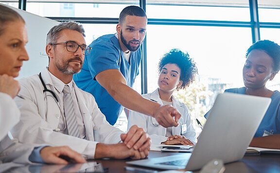 Eine Gruppe von fünf Fachleuten in medizinischer Kleidung bespricht Informationen auf einem Laptop. Ein Mann im blauen Oberteil weist auf den Bildschirm, während die anderen aufmerksam zuhören. Die Atmosphäre ist konzentriert und kollaborativ.