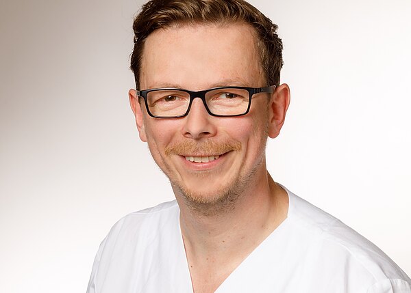 Porträt eines lächelnden Mannes mit kurzer Frisur und schwarzer Brille, in weißer medizinischer Kleidung, vor neutralem Hintergrund.