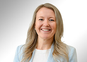 Professionelles Porträt einer lächelnden Frau mit langen, blonden Haaren in hellblauem Blazer und weißem Oberteil vor neutral hellem Hintergrund.