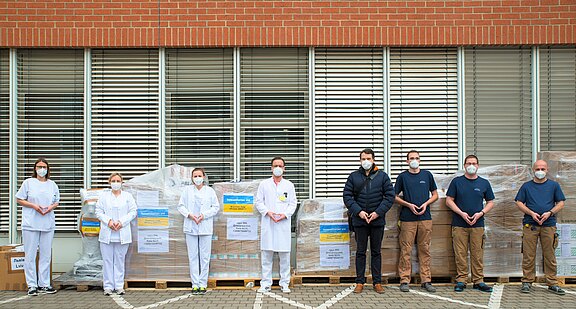 Eine Reihe von Menschen, teils in weißer Klinikbekleidung, alle mit Masken, steht vor mehreren in Folie verpackten Paletten voller Kisten. Aufkleber in Blau und Gelb weisen auf eine Hilfslieferung hin. Die Gruppe präsentiert die gesammelten medizinischen Materialien vor einem Gebäude.