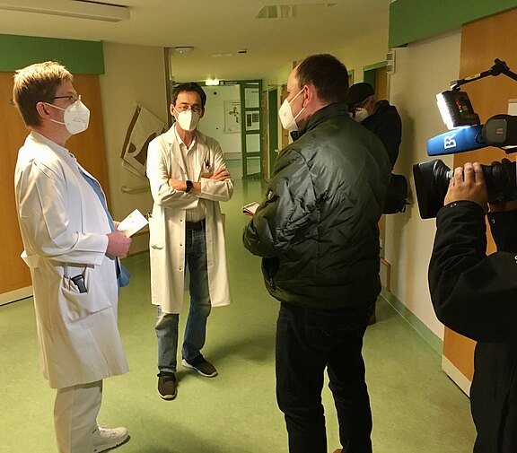 Im Krankenhausflur stehen zwei Ärztinnen/Ärzte mit Schutzmasken einem Reporter gegenüber; eine TV-Kamera zeichnet das Gespräch auf.