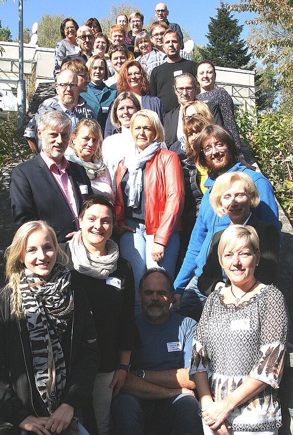 Großes Gruppenfoto: Rund 30 Erwachsene stehen dicht beisammen auf einer Außentreppe und schauen lächelnd in die Kamera. Sonniger Tag, informelle Kleidung. Der Fokus liegt auf der Gruppe, die für ein Foto posiert.