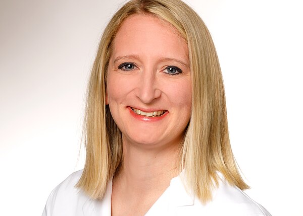 Porträt einer lächelnden erwachsenen Person mit schulterlangem blondem Haar in weißem Kittel, frontal zur Kamera vor hellem, neutralem Hintergrund.