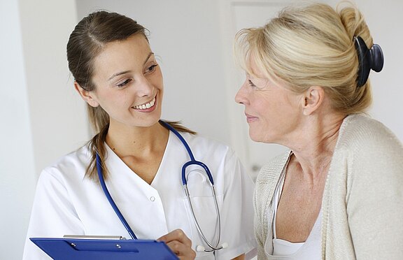 Smiling healthcare professional with a stethoscope holds a clipboard while talking with an older woman patient, suggesting a friendly medical consultation in a bright clinic.