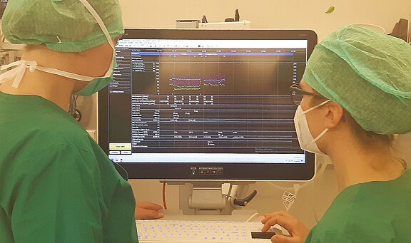 Zwei medizinisches Fachkräfte in grüner Kleidung stehen vor einem Computerbildschirm. Der Bildschirm zeigt eine Analyse mit Grafiken und Zahlen, die vermutlich medizinische Daten darstellen. Beide Personen tragen Masken und Hauben, was auf eine sterile Umgebung hinweist.