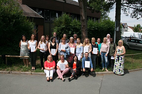 Gruppenfoto von etwa zwei Dutzend Erwachsenen, die im Freien vor einem Gebäude stehen. Viele halten Urkunden und lächeln in die Kamera – offenbar nach einer Abschluss- oder Zertifikatsverleihung.