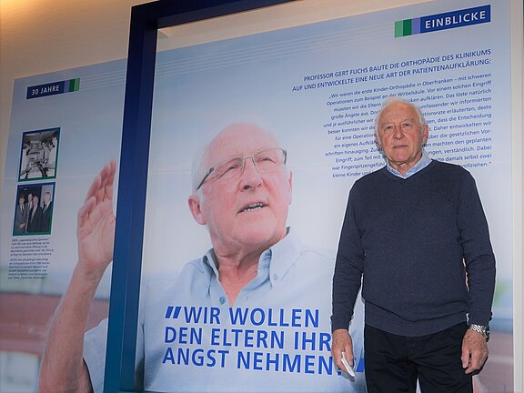 Älterer Mann steht vor einer großen Wand mit seinem Porträt und dem Zitat: „Wir wollen den Eltern ihre Angst nehmen.“ Die Ausstellung wirkt klinisch und würdigt langjährige Arbeit in der Orthopädie und Patientenaufklärung.