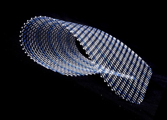 Ein metallisches Netzmaterial ist in einer geschwungenen Form dargestellt. Es zeigt eine Mischung aus silbernen und blauen Fäden, die sich spiralförmig anordnen. Der Hintergrund ist schwarz, was die Struktur des Materials hervorhebt.
