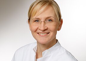 Das Bild zeigt eine lächelnde Frau mit blonden Haaren, die eine medizinische Fachjacke trägt. Sie hat eine Brille und steht vor einem hellen, einfarbigen Hintergrund. Ihr Ausdruck vermittelt Freundlichkeit und Professionalität.