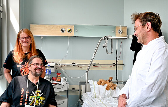 In einem Krankenhauszimmer unterhält sich ein lächelnder Arzt im weißen Kittel mit einem ebenso lachenden Patienten; neben ihm steht eine Frau, die zuschaut. Neben dem Bett mit Kissen und Teddybär wirkt die Szene freundlich und zugewandt.