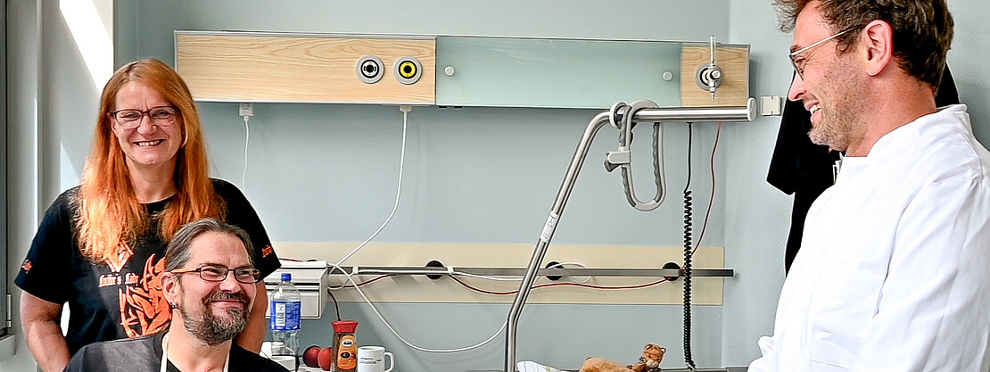 In einem Krankenhauszimmer unterhält sich ein lächelnder Arzt im weißen Kittel mit einem ebenso lachenden Patienten; neben ihm steht eine Frau, die zuschaut. Neben dem Bett mit Kissen und Teddybär wirkt die Szene freundlich und zugewandt.
