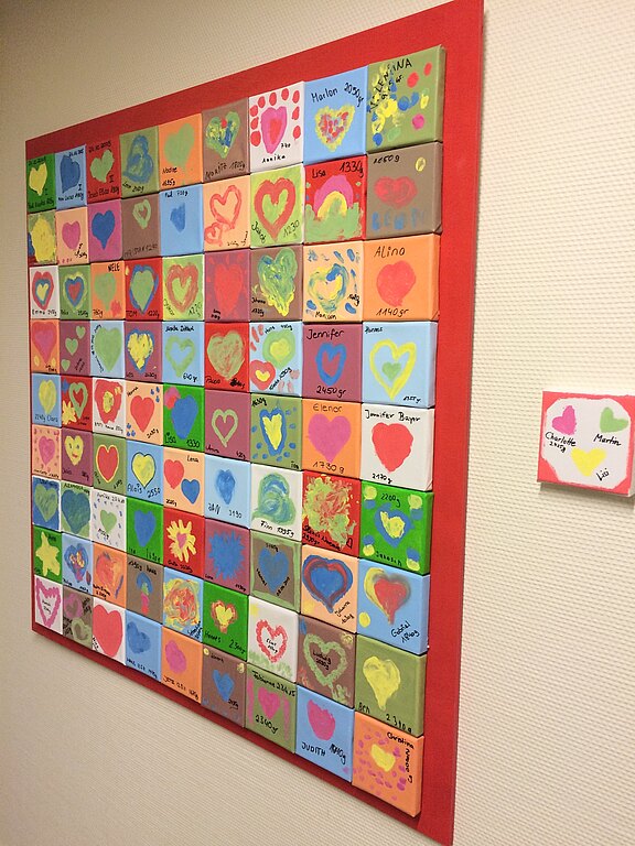 Buntes Wandbild aus vielen kleinen, verschiedenfarbigen Quadraten. In jedem Feld einfache, kindliche Motive, vor allem Herzen, teils mit Vornamen signiert. Insgesamt entsteht ein fröhliches Mosaik.
