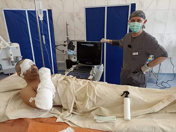 Ein verletzter Patient liegt in einem Krankenhausbett, Augen und rechter Arm stark verbunden. Neben ihm steht eine maskierte Pflegekraft vor einem Ultraschall- bzw. Monitorgerät. Die Szene zeigt medizinische Versorgung nach einer Verletzung oder Operation in einem Klinikraum.