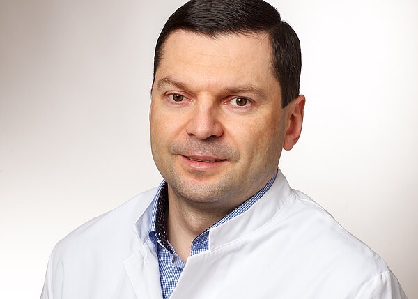 Porträt eines erwachsenen Mannes in weißem Kittel vor neutralem Hintergrund, leicht lächelnd, Blick zur Kamera.