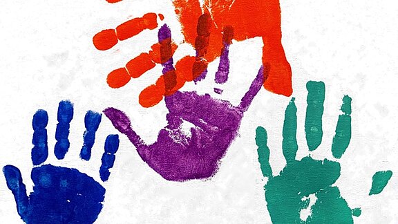 Vier bunte Handabdrücke auf weißem Hintergrund: blau links unten, grün rechts unten, lila mittig, darüber rot, teilweise überlappend. Eindruck von Kinder-Malerei und Vielfalt.