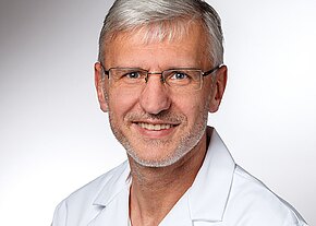 Porträt eines lächelnden Mannes mit grauen Haaren und Brille, im weißen Kittel, vor neutralem Hintergrund. Kopf und Schultern sind sichtbar.