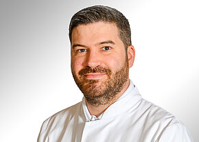 Porträt eines Mannes mit kurzem dunklem Haar und Bart, freundlich lächelnd, in weißem Kittel vor neutralem Hintergrund.