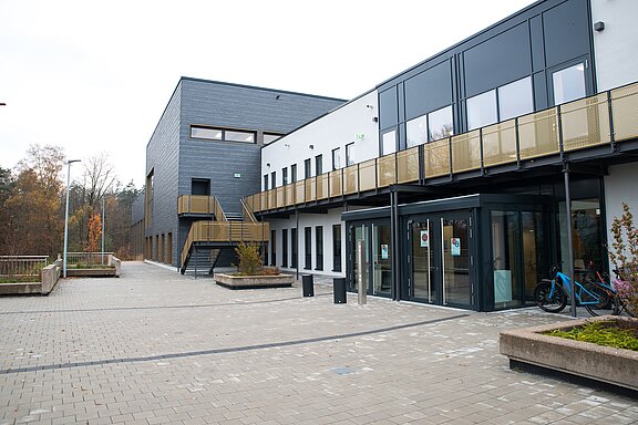 Moderner, zweigeschossiger Gebäudekomplex mit viel Glas und einem breiten, gepflasterten Vorplatz. Eine Außentreppe führt ins Obergeschoss, entlang der Fassade verläuft ein geländerter Laufsteg. Vor dem Eingang stehen Fahrradständer. Im Hintergrund Bäume, bedeckter Himmel.