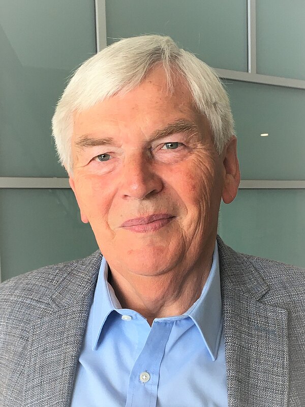 Porträt eines älteren Mannes mit kurzen grauen Haaren und freundlichem Ausdruck. Er trägt ein blaues Hemd und graues Sakko vor neutralem Hintergrund.