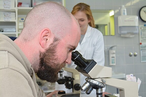 Ein Mann mit einem kurzen Haarschnitt und Bart schaut konzentriert durch ein Mikroskop. Im Hintergrund steht eine blonde Frau in einem weißen Kittel und beobachtet ihn. Die Szene spielt in einem Labor.