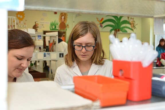 Zwei Frauen in Laborkitteln arbeiten in einem Labor. Eine Frau mit Brille schaut konzentriert auf ihre Aufgabe, während die andere leicht lächelt und ebenfalls beschäftigt ist. Im Vordergrund sind Labormaterialien zu sehen. Die Umgebung wirkt freundlich und einladend.