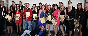 Eine Gruppe von Personen steht zusammen und hält Urkunden sowie Rosen in den Händen. Sie scheinen an einer Feier teilzunehmen, möglicherweise einer Abschlusszeremonie. Einige Teilnehmer lächeln und posieren freundlich für das Foto.