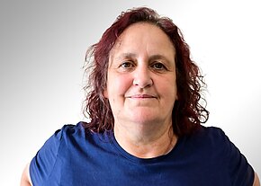 Susanne Dietze Porträt einer Frau mittleren Alters mit lockigem, rötlich-braunem Haar, Blick in die Kamera, dezentes Lächeln. Sie trägt ein dunkelblaues T‑Shirt. Heller, neutraler Hintergrund.