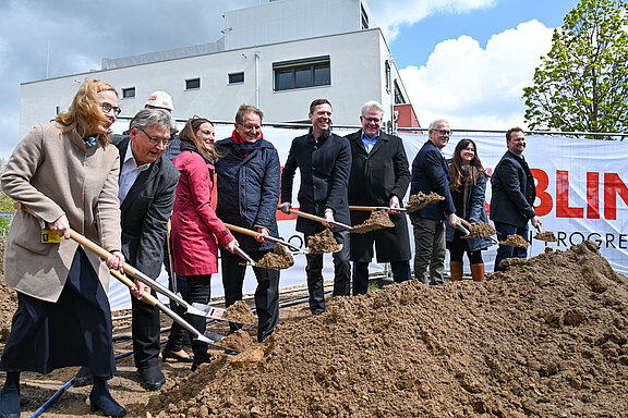 Eine Gruppe von Menschen steht an einer Baugrube und hält Schaufeln in der Hand. Sie sind bereit, den ersten Spatenstich zu setzen. Im Hintergrund ist ein Banner mit dem Wort "BAUBEGINN" sichtbar. Es ist ein sonniger Tag mit etwas bewölktem Himmel.
