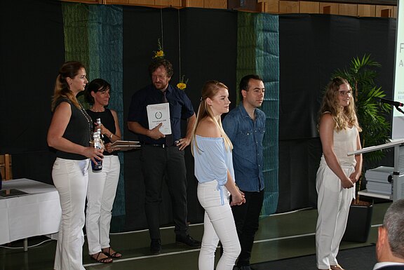 Mehrere junge Leute stehen auf einer Bühne und blicken zum Rednerpult. Hinter ihnen halten Erwachsene Urkunden bereit. Die Szene wirkt wie eine Schul- oder Abschlussfeier mit Preisverleihung.