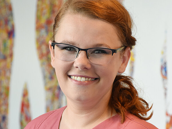 Porträt einer lächelnden Frau mit Brille in rosa Pflegeuniform, die direkt in die Kamera schaut. Sie trägt ein Namensschild; der Hintergrund ist unscharf.