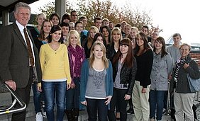 Eine Gruppe von etwa 30 Schülern und Lehrern steht draußen auf einer Treppe vor einem Gebäude. Einige der Schüler lächeln in die Kamera, während andere eine ernstere Miene haben. Im Vordergrund steht ein Lehrer in Anzug mit einer Krawatte. Herbstliche Bäume sind im Hintergrund sichtbar.