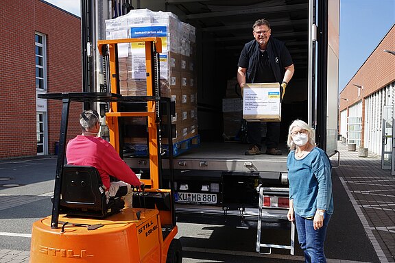 Ein Gabelstapler lädt Paletten in einen Lkw, während zwei Helfer Kartons mit der Aufschrift „Humanitäre Hilfe“ verladen.