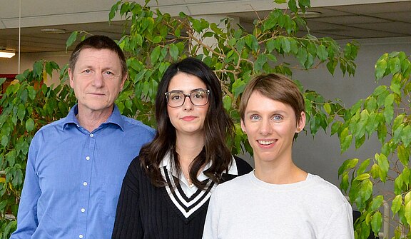 Drei Personen stehen nebeneinander und blicken in die Kamera. Links ein Mann im blauen Hemd, daneben zwei Frauen; die mittlere trägt Brille und schwarzen Pullover, die rechte ein helles Oberteil. Heller Innenraum mit grünen Pflanzen im Hintergrund. Lockeres, freundliches Gruppenporträt.