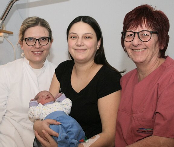Eine frischgebackene Mutter hält ihr schlafendes Neugeborenes im Arm. Neben ihr sitzen zwei lächelnde Medizinerinnen in Klinikkleidung. Die Szene vermittelt Freude nach der Geburt und fürsorgliche Betreuung.