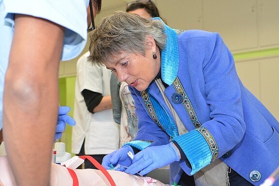 Eine Person mit grauen Haaren und blauer Jacke führt eine medizinische Handlung durch, während sie auf ein simuliertes Patientenmodell schaut. Sie trägt Handschuhe und konzentriert sich auf den Vorgang. Im Hintergrund sind weitere Personen in weißen Oberteilen zu sehen.