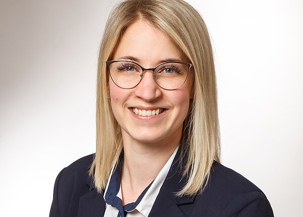 Porträtfoto: lächelnde Frau mit schulterlangem blondem Haar und Brille, in dunkelblauem Blazer und weißer Bluse. Heller Hintergrund; professionelles Business-Porträt.