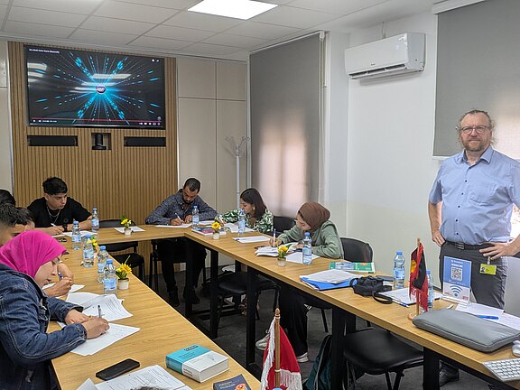 Eine Gruppe von vier Personen sitzt an einem Tisch und schreibt Notizen, während ein Mann im Hintergrund steht. Auf einem Bildschirm im Raum sind visuelle Elemente zu sehen. Es sind Wasserflaschen und kleine Pflanzen auf dem Tisch platziert.