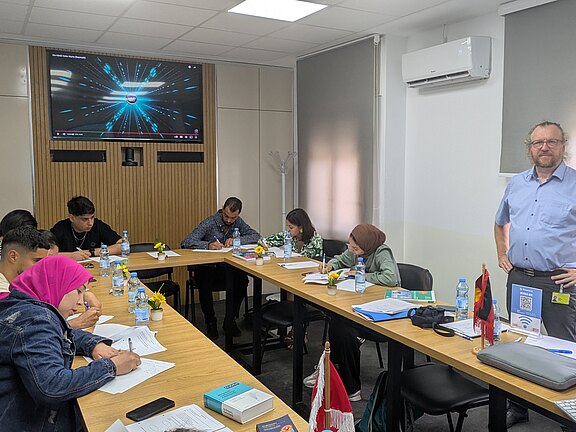 Eine Gruppe von vier Personen sitzt an einem Tisch und schreibt Notizen, während ein Mann im Hintergrund steht. Auf einem Bildschirm im Raum sind visuelle Elemente zu sehen. Es sind Wasserflaschen und kleine Pflanzen auf dem Tisch platziert.