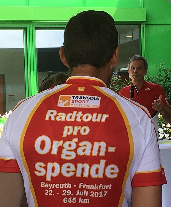 Rückenansicht eines Radfahrers im Trikot mit Aufdruck: „Radtour pro Organspende“. Oben Logo „Transdia Sport Deutschland“. Darunter Route Bayreuth–Frankfurt, 22.–29. Juli 2017, 645 km. Im Hintergrund spricht eine Person. Motiv wirbt für Organspende.