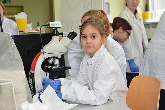 Ein Mädchen mit braunen Haaren sitzt an einem Tisch in einem Labor, trägt einen weißen Laborkittel und blaue Handschuhe. Vor ihr steht ein Mikroskop. Im Hintergrund sind weitere Personen in Laborkitteln zu sehen, die ebenfalls im Labor arbeiten.