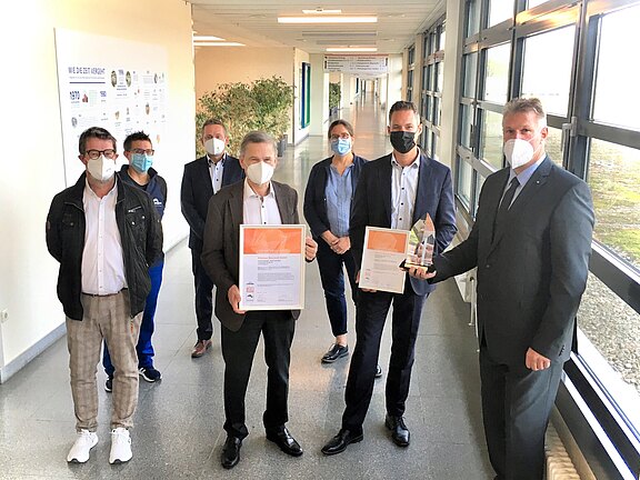 Gruppe von sieben Personen in einem Flur, alle mit Masken. Zwei Personen vorn halten Urkunden, eine weitere hält einen gläsernen Preis. Es wirkt wie eine kleine Preisverleihung in einem Büro- oder Klinikgebäude.