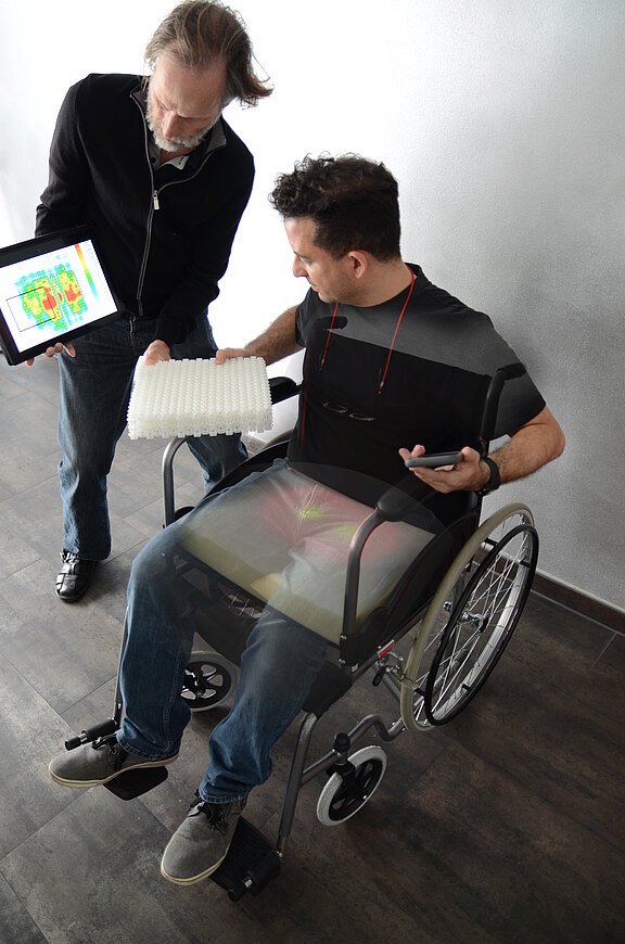 Zwei Personen prüfen die Sitzposition eines Rollstuhlfahrers. Der Stehende zeigt auf einem Tablet eine Druckverteilungs-Heatmap und hält ein geriffeltes Sitzkissen. Ziel ist die Anpassung der Sitzunterlage zur Druckentlastung und Komfortverbesserung.
