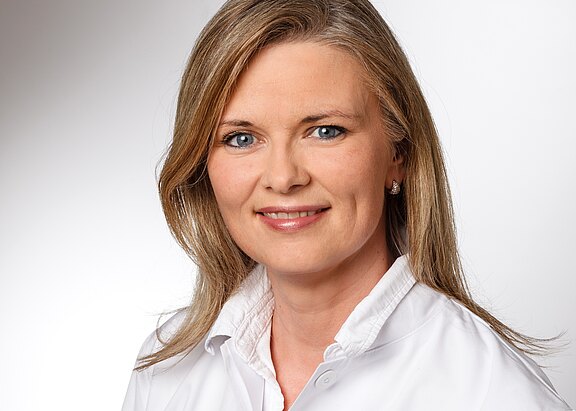 Porträt einer lächelnden Frau mit schulterlangen blonden Haaren in weißem Kittel, frontal zur Kamera vor hellem, neutralem Hintergrund.