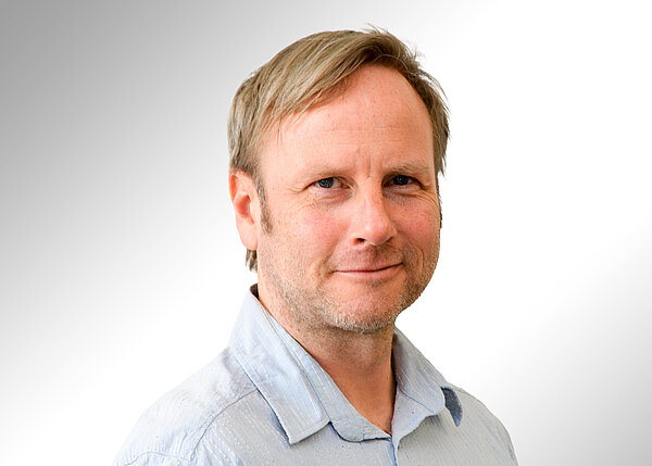 Porträt eines freundlich blickenden Mannes mittleren Alters mit hellblonden Haaren und leichten Bartstoppeln, in hellblauem Hemd, vor neutralem, hellgrau-weißem Hintergrund.