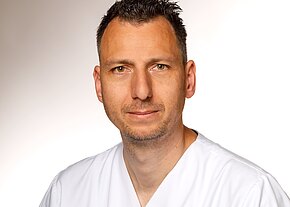 Station 34 Porträt eines Mannes mittleren Alters in weißer medizinischer Kleidung vor neutralem Hintergrund. Er blickt ruhig in die Kamera.